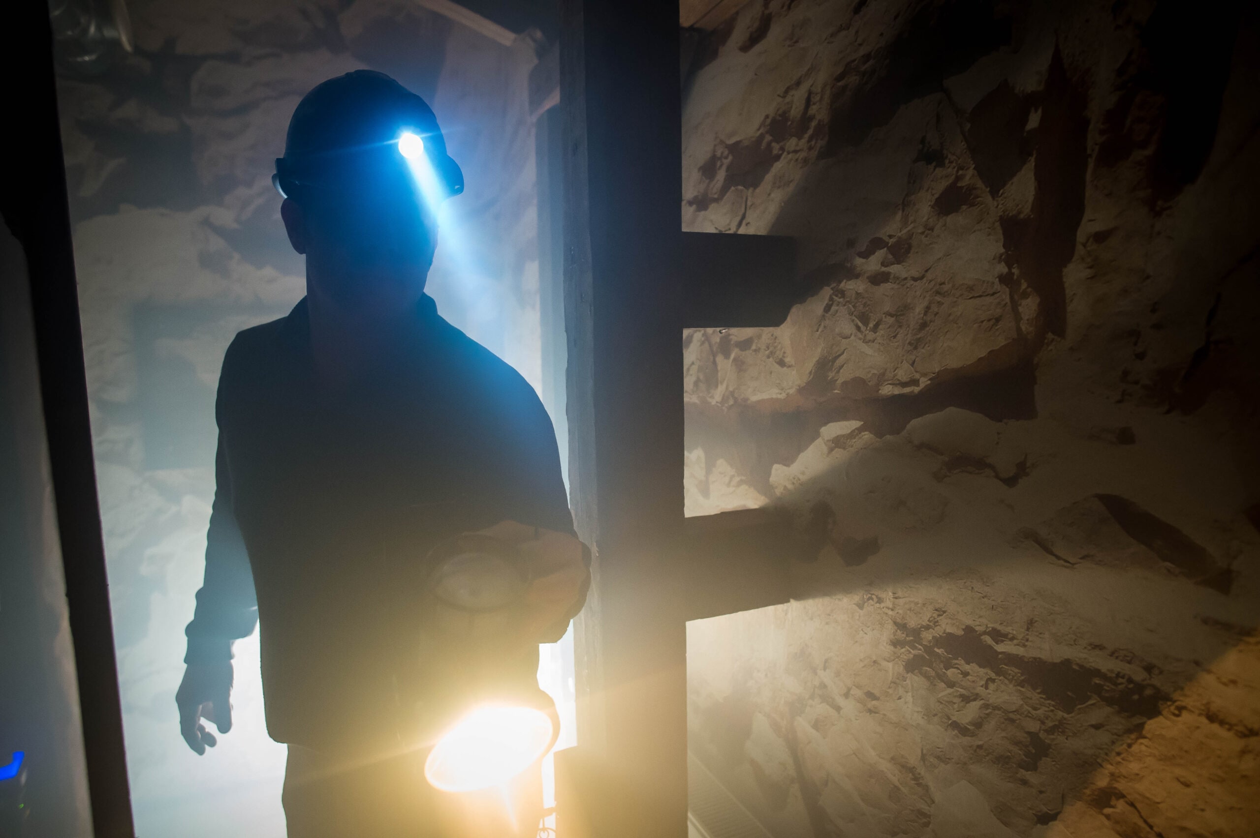 Ein Mann in Arbeitskleidung mit Helm und Stirnlampe steht in einem staubigen, undeutlich beleuchteten Raum, umgeben von unverputzten Steinwänden.